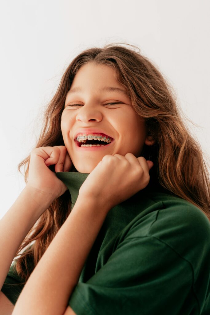 Joyful portrait of a teenage girl in a green shirt showing braces and a bright smile against a white background.